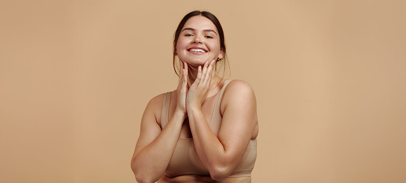 A woman smiling and touching her face with both hands, standing against a plain beige background. She is wearing a beige sports bra.