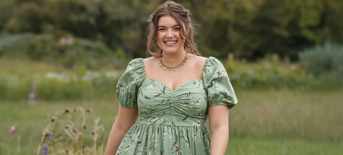 A smiling woman with long brown hair stands outdoors in a grassy field, wearing a light green floral dress with puffed sleeves and a gold necklace.