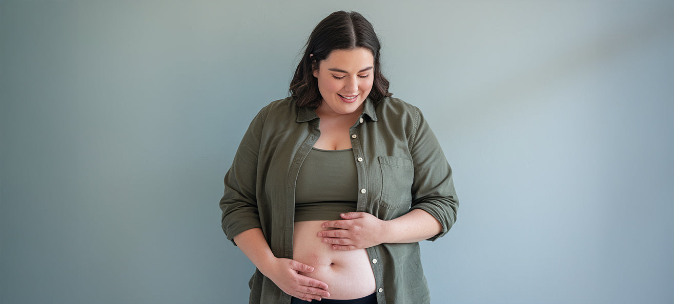 A woman standing against a plain light blue wall, smiling and looking down while gently holding her bare stomach with both hands. She is wearing a green open button-up shirt over a matching tank top.