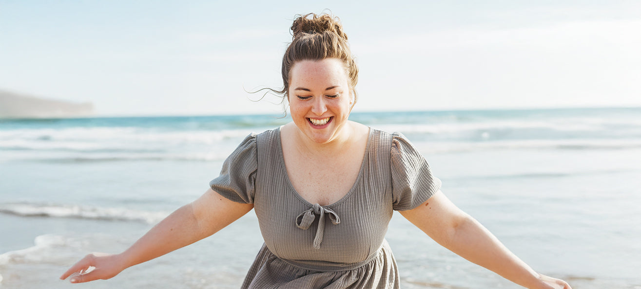 A joyful woman smiling with her arms outstretched while standing on a beach in a short-sleeve dress. The ocean waves and blue sky are in the background.