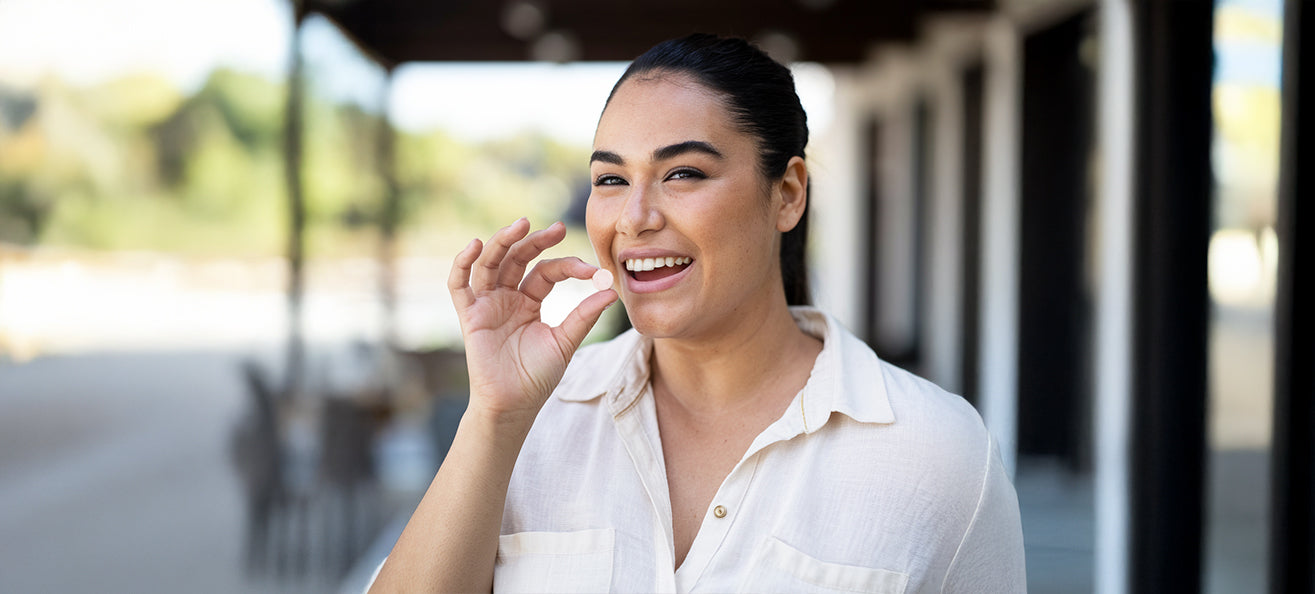 A Latina woman smiling outdoors while holding a round supplement tablet near her mouth. She appears happy and confident, wearing a light-colored button-up shirt, with a blurred background of trees and buildings.