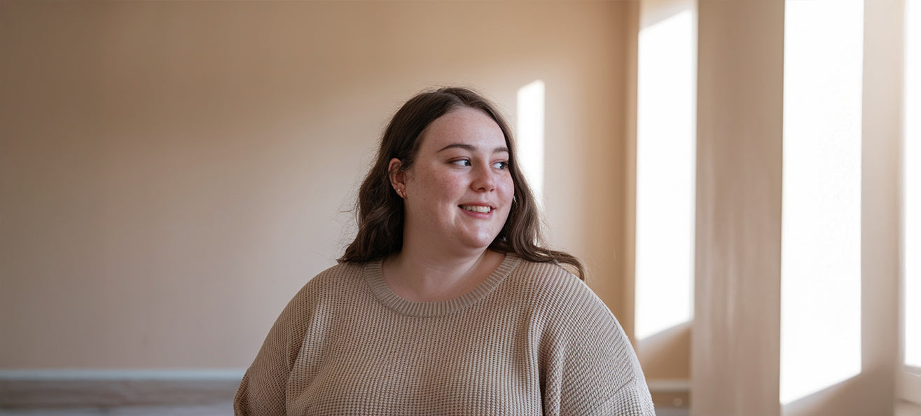A woman with long brown hair smiles softly while looking to the side, wearing a beige sweater in a bright sunlit room.